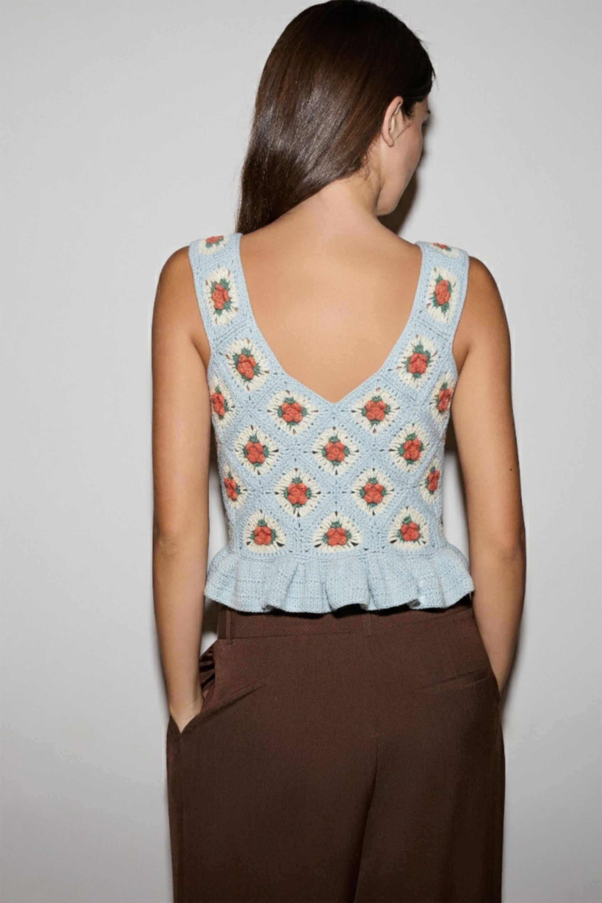 Woman wearing a sleeveless floral top with ruffled details and brown pants on a plain background