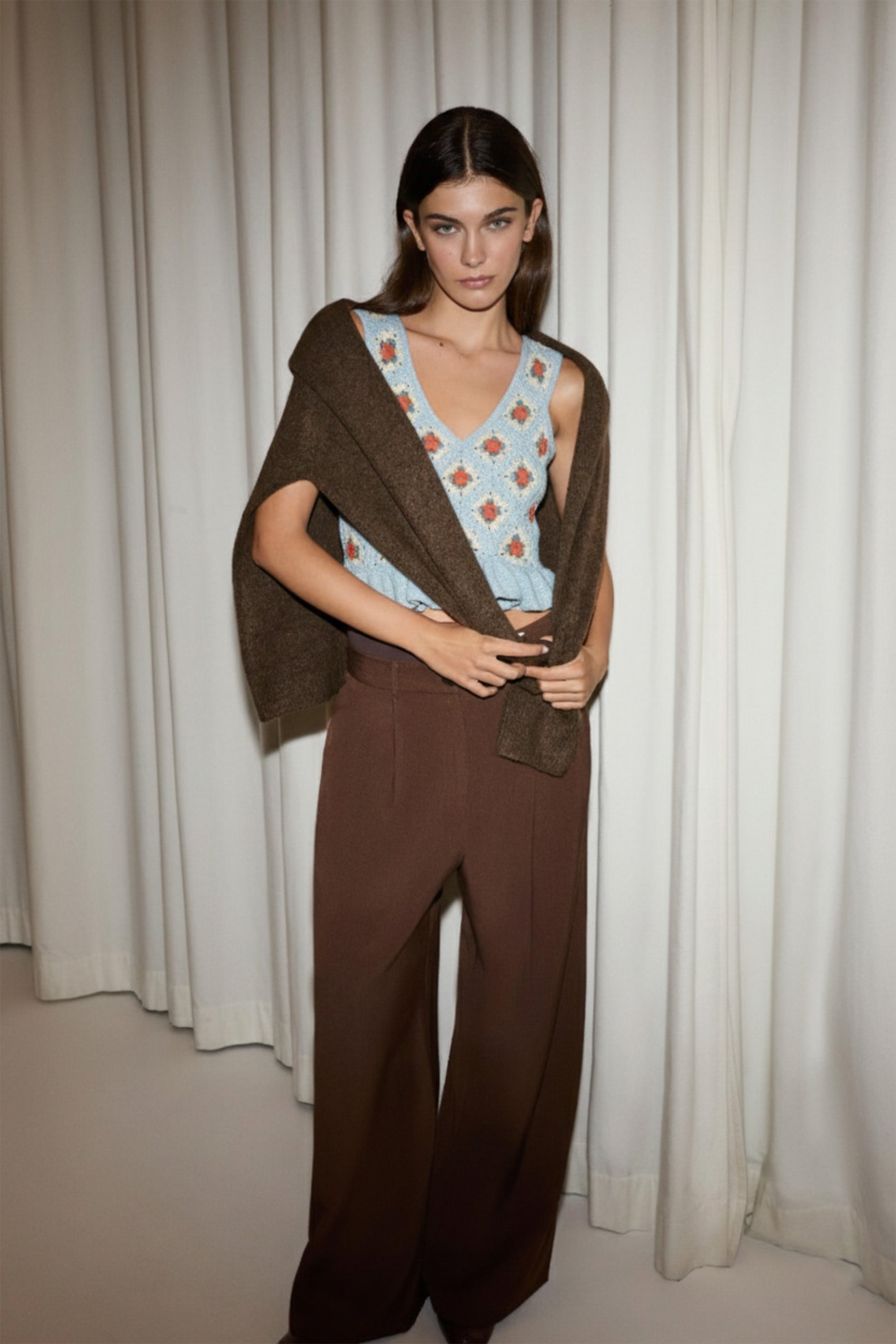 Woman wearing a brown poncho over a light blue top with a geometric pattern and brown pants against a white curtain background.